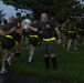 A large group of military personnel, appearing to be U.S. Marines based on their olive drab PT (physical training) uniforms, are conducting a formation run in an outdoor area. The participants are wearing matching olive green t-shirts and shorts with high-visibility yellow/neon safety belts. A senior figure, identifiable by black knee-high socks and a unit insignia on his shirt, appears to be directing or guiding runners near the front.