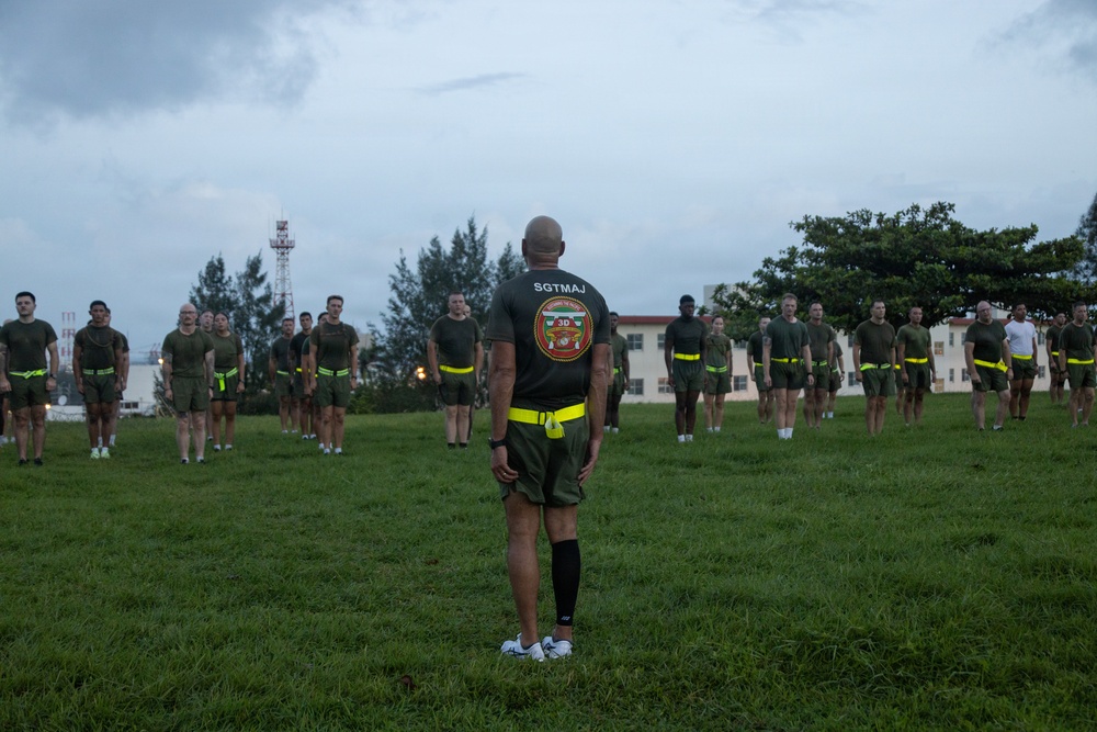 A Sergeant Major (SGTMAJ) of what appears to be the 3rd Marine Division stands with his back to the camera, facing a formation of military personnel on a grass field. The troops are wearing olive drab PT (physical training) shorts and shirts with neon yellow/green belts, suggesting a physical training session. The setting appears to be a military base, likely in a Pacific location given the tropical vegetation and architecture.