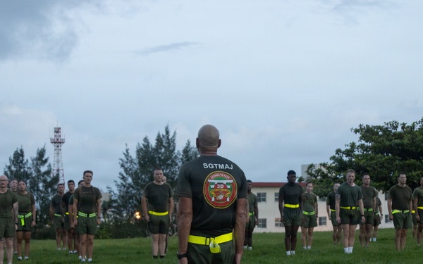 A Sergeant Major (SGTMAJ) of what appears to be the 3rd Marine Division stands with his back to the camera, facing a formation of military personnel on a grass field. The troops are wearing olive drab PT (physical training) shorts and shirts with neon yellow/green belts, suggesting a physical training session. The setting appears to be a military base, likely in a Pacific location given the tropical vegetation and architecture.
