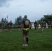 A Sergeant Major (SGTMAJ) of what appears to be the 3rd Marine Division stands with his back to the camera, facing a formation of military personnel on a grass field. The troops are wearing olive drab PT (physical training) shorts and shirts with neon yellow/green belts, suggesting a physical training session. The setting appears to be a military base, likely in a Pacific location given the tropical vegetation and architecture.