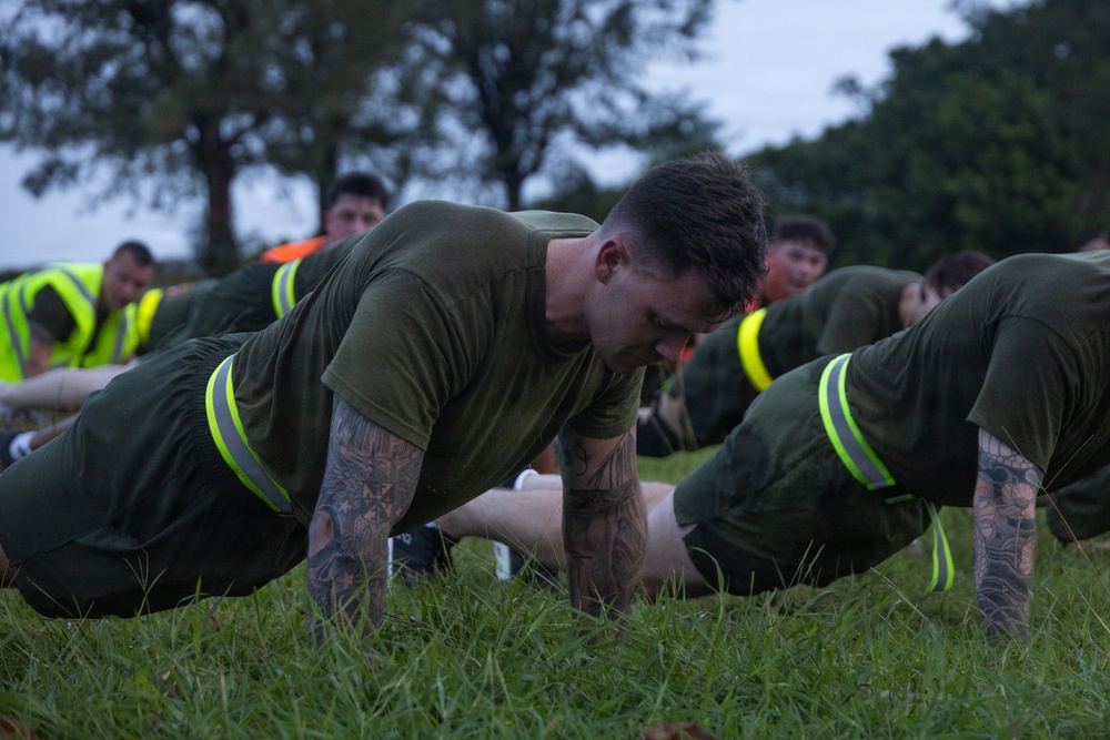 A group of military personnel, likely U.S. Marines based on their olive drab PT uniforms, are performing push-ups on a grass field during what appears to be an early morning physical training session. The foreground subject is a heavily tattooed male in the up position of a push-up. Supervisors or safety personnel wearing high-visibility yellow vests are visible in the background.