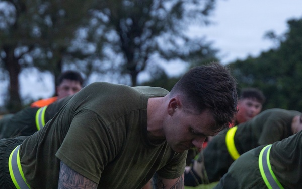 A group of military personnel, likely U.S. Marines based on their olive drab PT uniforms, are performing push-ups on a grass field during what appears to be an early morning physical training session. The foreground subject is a heavily tattooed male in the up position of a push-up. Supervisors or safety personnel wearing high-visibility yellow vests are visible in the background.