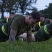 A group of military personnel, likely U.S. Marines based on their olive drab PT uniforms, are performing push-ups on a grass field during what appears to be an early morning physical training session. The foreground subject is a heavily tattooed male in the up position of a push-up. Supervisors or safety personnel wearing high-visibility yellow vests are visible in the background.
