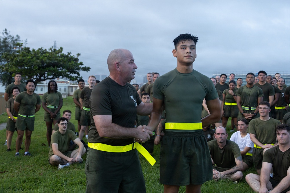 A military or law enforcement physical training session is taking place outdoors, likely at a base or facility near an urban area. An older, bald instructor wearing a yellow high-visibility belt is shaking hands with or presenting recognition to a younger service member who is also wearing a yellow reflective belt. A large group of uniformed personnel in olive drab PT gear are gathered as an audience, some seated on the grass and others standing.