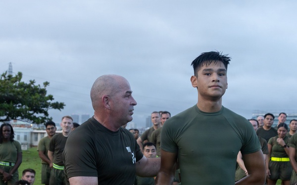 A military or law enforcement physical training session is taking place outdoors, likely at a base or facility near an urban area. An older, bald instructor wearing a yellow high-visibility belt is shaking hands with or presenting recognition to a younger service member who is also wearing a yellow reflective belt. A large group of uniformed personnel in olive drab PT gear are gathered as an audience, some seated on the grass and others standing.