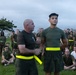 A military or law enforcement physical training session is taking place outdoors, likely at a base or facility near an urban area. An older, bald instructor wearing a yellow high-visibility belt is shaking hands with or presenting recognition to a younger service member who is also wearing a yellow reflective belt. A large group of uniformed personnel in olive drab PT gear are gathered as an audience, some seated on the grass and others standing.