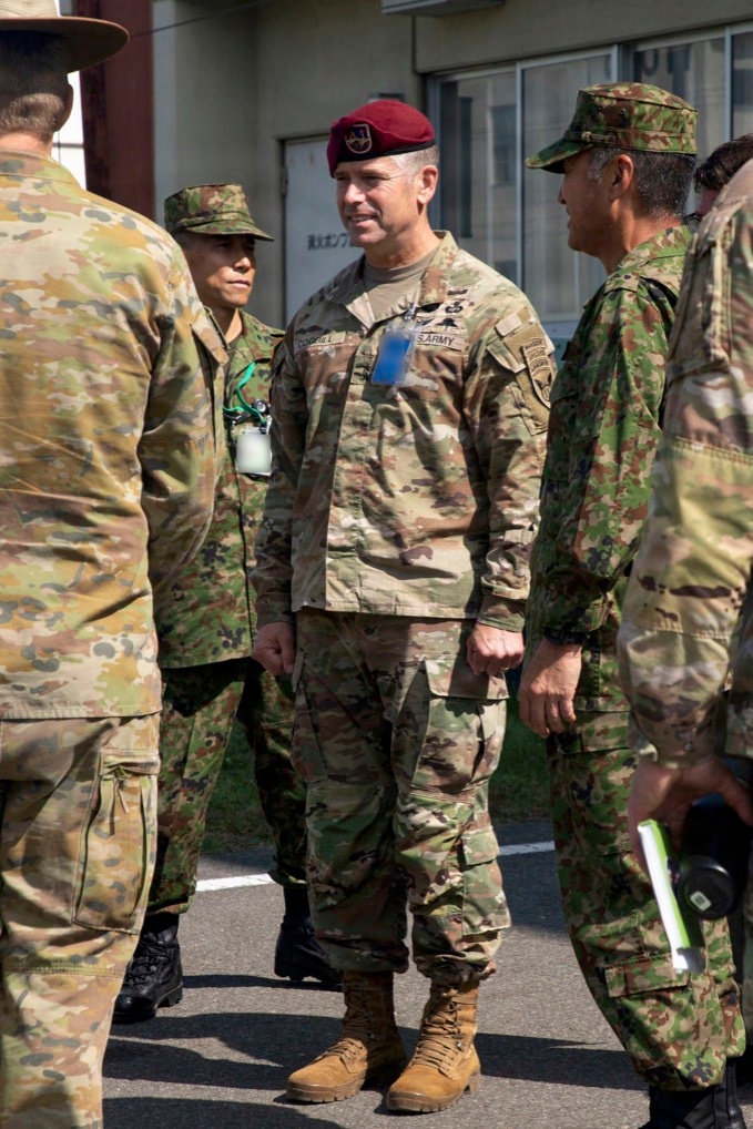 A U.S. Army soldier wearing a maroon beret and OCP (Operational Camouflage Pattern) uniform is standing among a group of military personnel, some of whom appear to be wearing Japanese Ground Self-Defense Force (JGSDF) camouflage uniforms. The central figure has a name tape reading 'COGHILL' and 'U.S. ARMY' visible on their uniform, along with rank insignia and unit patches. The setting appears to be an outdoor area near a building with Japanese text visible, suggesting a joint military exercise or visit.