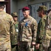 A U.S. Army soldier wearing a maroon beret and OCP (Operational Camouflage Pattern) uniform is standing among a group of military personnel, some of whom appear to be wearing Japanese Ground Self-Defense Force (JGSDF) camouflage uniforms. The central figure has a name tape reading 'COGHILL' and 'U.S. ARMY' visible on their uniform, along with rank insignia and unit patches. The setting appears to be an outdoor area near a building with Japanese text visible, suggesting a joint military exercise or visit.