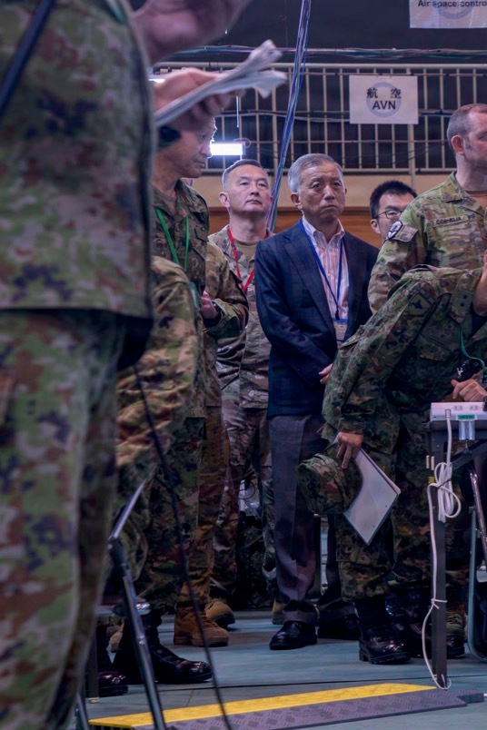 The image shows a multinational military briefing or command post exercise, likely a joint US-Japan military operation based on visible signage and uniform types. Personnel in Japanese Ground Self-Defense Force (JGSDF) camouflage uniforms, US Army MultiCam uniforms, and civilian attire are gathered attentively, apparently observing a presentation or briefing. A sign in the background displays Japanese characters (航空) alongside the acronym 'AVN' (Aviation), suggesting an aviation-related operations context.