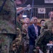 The image shows a multinational military briefing or command post exercise, likely a joint US-Japan military operation based on visible signage and uniform types. Personnel in Japanese Ground Self-Defense Force (JGSDF) camouflage uniforms, US Army MultiCam uniforms, and civilian attire are gathered attentively, apparently observing a presentation or briefing. A sign in the background displays Japanese characters (航空) alongside the acronym 'AVN' (Aviation), suggesting an aviation-related operations context.