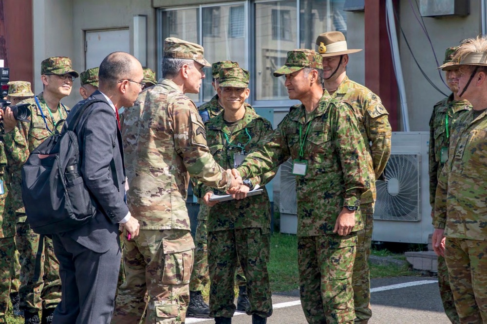 A multinational military gathering showing personnel from at least three different armed forces engaged in formal greetings. A central handshake is taking place between officers, suggesting an official meeting or ceremony. A civilian in a business suit is also present among the military personnel.