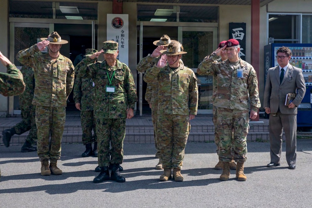A group of military personnel from multiple nations are rendering salutes in front of a building with Japanese signage, suggesting a formal military ceremony or official visit. The group includes soldiers wearing Australian Army slouch hats, Japanese Ground Self-Defense Force uniforms, and US Army uniforms including one with a maroon beret. A civilian in a suit stands to the right, likely serving as an interpreter or liaison.