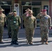 A group of military personnel from multiple nations are rendering salutes in front of a building with Japanese signage, suggesting a formal military ceremony or official visit. The group includes soldiers wearing Australian Army slouch hats, Japanese Ground Self-Defense Force uniforms, and US Army uniforms including one with a maroon beret. A civilian in a suit stands to the right, likely serving as an interpreter or liaison.