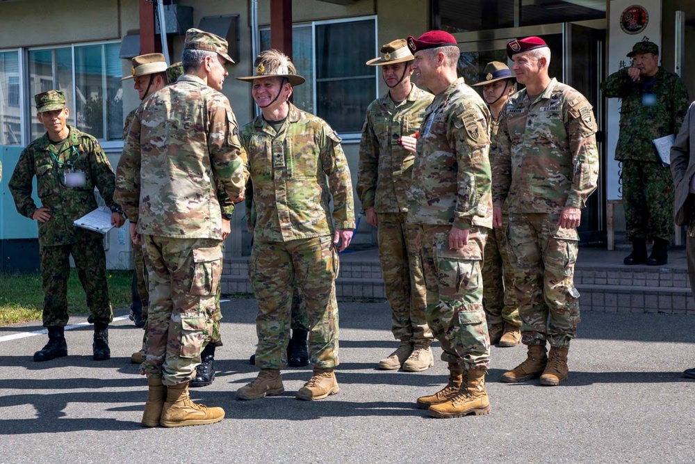 A group of multinational military personnel are gathered outdoors in front of a building, appearing to be engaged in informal conversation. The group includes soldiers wearing different national uniforms and headgear, suggesting a joint military exercise or official meeting. The setting appears to be a military installation, likely in Japan based on visible Japanese signage and Japanese Ground Self-Defense Force personnel present.