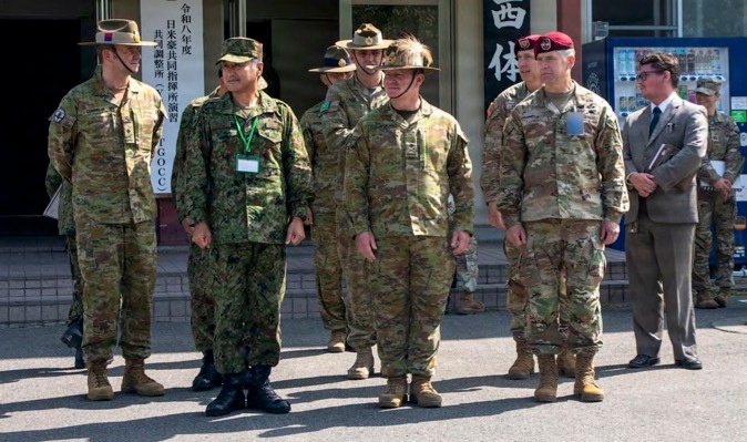 A group of military personnel from multiple nations are assembled outdoors in front of a building with Japanese signage. The scene appears to be a multinational military exercise or coordination event, likely in Japan, based on the visible kanji text referencing a joint command post exercise (JTGOCC). Personnel are wearing distinct uniforms representing at least three different military forces, including Japanese Ground Self-Defense Force, Australian Army, and United States Army.