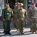 A group of military personnel from multiple nations are assembled outdoors in front of a building with Japanese signage. The scene appears to be a multinational military exercise or coordination event, likely in Japan, based on the visible kanji text referencing a joint command post exercise (JTGOCC). Personnel are wearing distinct uniforms representing at least three different military forces, including Japanese Ground Self-Defense Force, Australian Army, and United States Army.