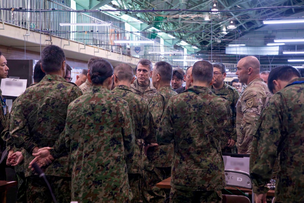 A group of military personnel from at least two different armed forces are gathered in what appears to be an indoor operations or briefing facility. The majority wear a distinctive green/brown woodland camouflage pattern consistent with Japan Ground Self-Defense Force (JGSDF) uniforms, while at least two individuals wear MultiCam/OCP pattern uniforms consistent with U.S. Army attire. The group appears to be engaged in a collaborative briefing or coordination meeting.