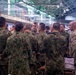 A group of military personnel from at least two different armed forces are gathered in what appears to be an indoor operations or briefing facility. The majority wear a distinctive green/brown woodland camouflage pattern consistent with Japan Ground Self-Defense Force (JGSDF) uniforms, while at least two individuals wear MultiCam/OCP pattern uniforms consistent with U.S. Army attire. The group appears to be engaged in a collaborative briefing or coordination meeting.