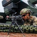 A U.S. Army soldier in full combat gear is positioned prone behind a light machine gun mounted on a bipod, aiming downrange during what appears to be a training exercise or demonstration. The soldier is wearing MultiCam (OCP) camouflage, a tactical helmet with accessories, and ballistic sunglasses. A second soldier and a large military vehicle or weapons system are visible in the background.