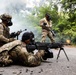 Military personnel in camouflage uniforms and gas masks are conducting a training exercise on a paved road surrounded by vegetation. Smoke and fire are visible in the background, consistent with a simulated combat or NBC (Nuclear, Biological, Chemical) training scenario. One soldier is in a prone firing position with a light machine gun on a bipod, while others provide security and one individual wearing a yellow safety vest appears to be an observer or evaluator.