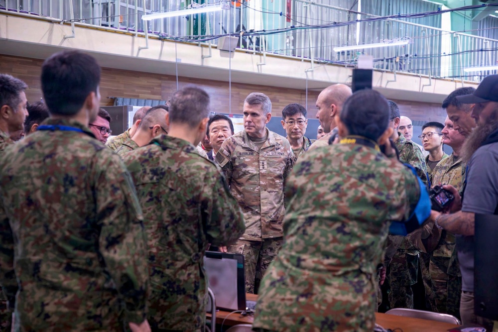 A group of military personnel from what appears to be the U.S. Army and Japanese Ground Self-Defense Force (JGSDF) are gathered in an indoor facility, likely during a joint briefing or coordination meeting. A central U.S. Army officer in Operational Camouflage Pattern (OCP) uniform with a name tape reading 'McFarlane' is visible, surrounded by personnel in both U.S. and Japanese camouflage uniforms. The setting suggests a joint military exercise or command visit.