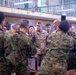 A group of military personnel from what appears to be the U.S. Army and Japanese Ground Self-Defense Force (JGSDF) are gathered in an indoor facility, likely during a joint briefing or coordination meeting. A central U.S. Army officer in Operational Camouflage Pattern (OCP) uniform with a name tape reading 'McFarlane' is visible, surrounded by personnel in both U.S. and Japanese camouflage uniforms. The setting suggests a joint military exercise or command visit.