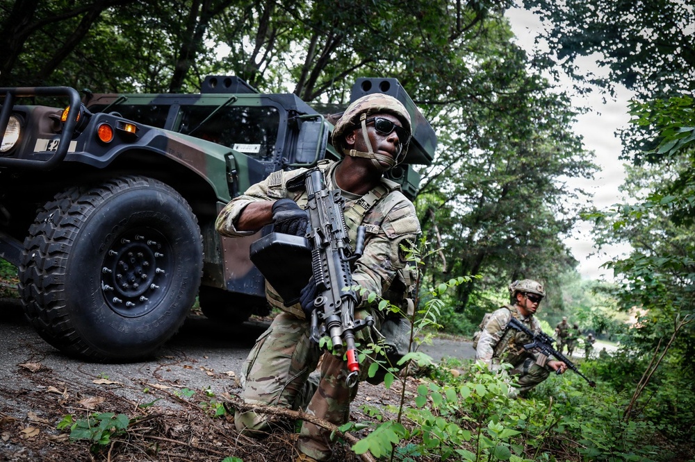 The image shows U.S. Army soldiers conducting a tactical field exercise in a wooded area alongside a military HMMWV (Humvee). The foreground soldier is in a kneeling security position holding an M4/M16-style assault rifle with an underslung grenade launcher attachment. Additional soldiers are visible in the background moving through the tree line.