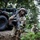 The image shows U.S. Army soldiers conducting a tactical field exercise in a wooded area alongside a military HMMWV (Humvee). The foreground soldier is in a kneeling security position holding an M4/M16-style assault rifle with an underslung grenade launcher attachment. Additional soldiers are visible in the background moving through the tree line.