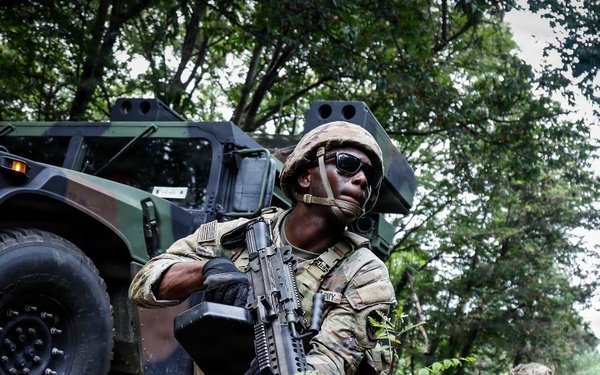 The image shows U.S. Army soldiers conducting a tactical field exercise in a wooded area alongside a military HMMWV (Humvee). The foreground soldier is in a kneeling security position holding an M4/M16-style assault rifle with an underslung grenade launcher attachment. Additional soldiers are visible in the background moving through the tree line.