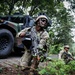 The image shows U.S. Army soldiers conducting a tactical field exercise in a wooded area alongside a military HMMWV (Humvee). The foreground soldier is in a kneeling security position holding an M4/M16-style assault rifle with an underslung grenade launcher attachment. Additional soldiers are visible in the background moving through the tree line.