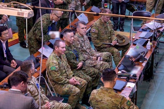 A multinational military command post or operations center showing personnel from multiple nations gathered around long tables equipped with laptops and communications equipment. The scene appears to be a joint military exercise or briefing, with personnel wearing various national uniforms including Australian Army (AUSCAM pattern), US Army (OCP/Multicam), and Japanese Ground Self-Defense Force uniforms. Civilian personnel in suits are also present, suggesting a high-level coordination or liaison event.