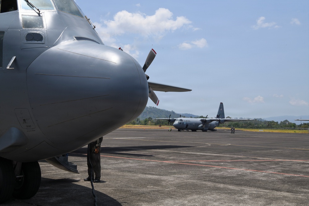 The image shows at least two military turboprop transport aircraft, consistent with C-130 Hercules variants, parked on a tarmac at what appears to be a tropical airfield. A ground crew member is visible standing beneath the nose of the foreground aircraft performing inspection or maintenance. A second aircraft is visible in the background with additional personnel nearby.