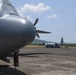 The image shows at least two military turboprop transport aircraft, consistent with C-130 Hercules variants, parked on a tarmac at what appears to be a tropical airfield. A ground crew member is visible standing beneath the nose of the foreground aircraft performing inspection or maintenance. A second aircraft is visible in the background with additional personnel nearby.