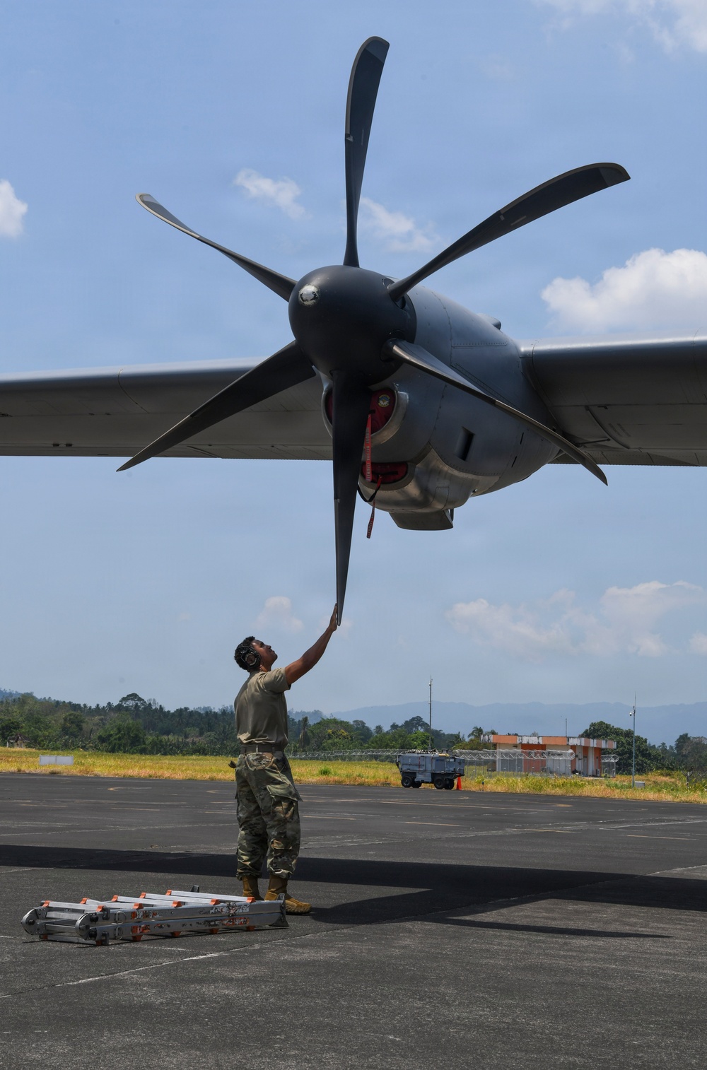 A military aircraft maintainer in camouflage uniform is performing a pre-flight or post-flight inspection on a large turboprop aircraft, manually checking or positioning one of the propeller blades. The aircraft appears to be a C-130 or similar military transport, featuring a large six-bladed propeller. The scene takes place on a tarmac at what appears to be a tropical or subtropical airfield, with lush green hills and a building visible in the background.