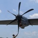 A military aircraft maintainer in camouflage uniform is performing a pre-flight or post-flight inspection on a large turboprop aircraft, manually checking or positioning one of the propeller blades. The aircraft appears to be a C-130 or similar military transport, featuring a large six-bladed propeller. The scene takes place on a tarmac at what appears to be a tropical or subtropical airfield, with lush green hills and a building visible in the background.