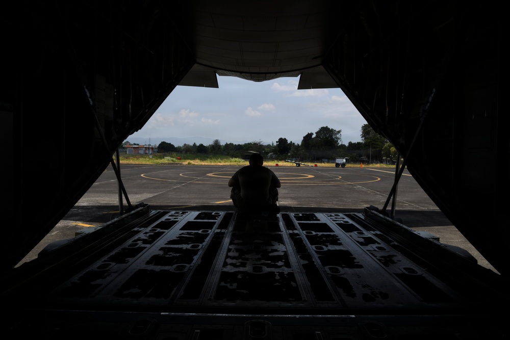 A silhouetted figure sits on the open rear cargo ramp of a military transport aircraft, looking out onto a tarmac or airfield. The perspective is from inside the aircraft's cargo hold, framing the exterior scene through the open ramp door. The airfield appears to be in a tropical or subtropical location based on the surrounding vegetation.