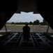A silhouetted figure sits on the open rear cargo ramp of a military transport aircraft, looking out onto a tarmac or airfield. The perspective is from inside the aircraft's cargo hold, framing the exterior scene through the open ramp door. The airfield appears to be in a tropical or subtropical location based on the surrounding vegetation.
