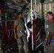 Four military personnel are standing inside the cargo hold of a large military transport aircraft, likely a C-130 or similar aircraft. They appear to be engaged in a briefing or inspection of the aircraft's interior equipment. The personnel are wearing a mix of OCP (Operational Camouflage Pattern) uniforms and tan t-shirts with military trousers.