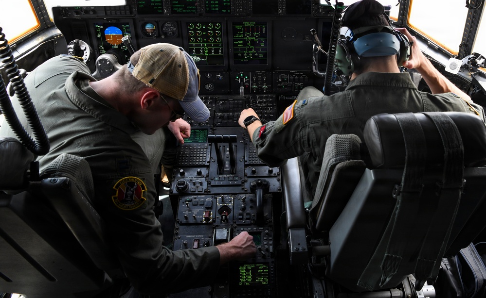 Two military aircrew members are seated in the cockpit of a large military aircraft, likely a C-130 Hercules or similar transport aircraft. The crew members are actively working with the flight controls and instrument panels, which display numerous green-lit digital readouts and analog gauges. One crew member wears a baseball cap and flight suit with a squadron patch, while the other wears a flight helmet with communications headset.
