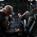 Two military aircrew members are seated in the cockpit of a large military aircraft, likely a C-130 Hercules or similar transport aircraft. The crew members are actively working with the flight controls and instrument panels, which display numerous green-lit digital readouts and analog gauges. One crew member wears a baseball cap and flight suit with a squadron patch, while the other wears a flight helmet with communications headset.