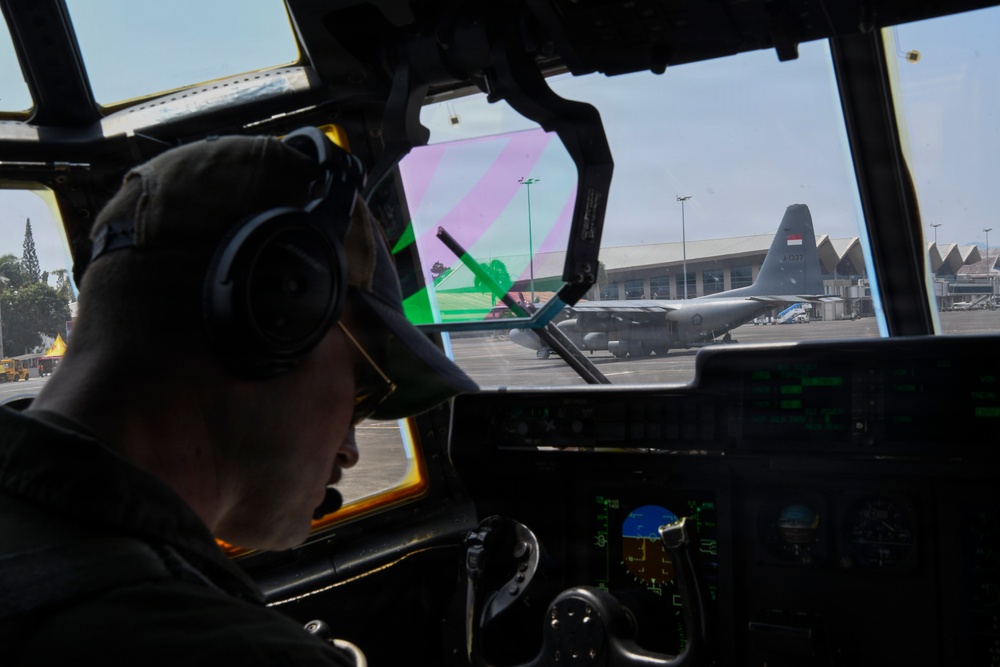A military pilot or crew member is seated in the cockpit of a military transport aircraft, viewed from behind. Through the windshield, a C-130 Hercules transport aircraft bearing what appears to be an Indonesian flag roundel is visible on the tarmac. The scene appears to be at a military or joint-use airport facility.