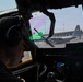 A military pilot or crew member is seated in the cockpit of a military transport aircraft, viewed from behind. Through the windshield, a C-130 Hercules transport aircraft bearing what appears to be an Indonesian flag roundel is visible on the tarmac. The scene appears to be at a military or joint-use airport facility.
