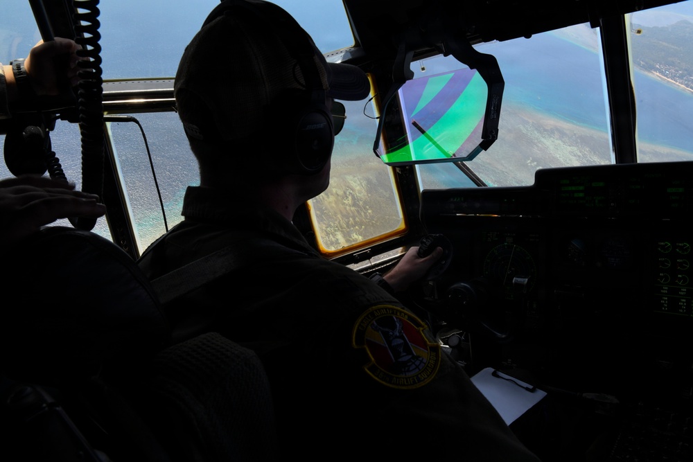 A military pilot is seated in the cockpit of a large aircraft, flying over a coastal area with visible reef and turquoise water below. The pilot is wearing a flight suit with a squadron patch, a headset, and a baseball cap. A colorful parachute or drogue chute is visible through the windscreen, suggesting an airdrop or aerial delivery operation may be in progress.