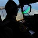 A military pilot is seated in the cockpit of a large aircraft, flying over a coastal area with visible reef and turquoise water below. The pilot is wearing a flight suit with a squadron patch, a headset, and a baseball cap. A colorful parachute or drogue chute is visible through the windscreen, suggesting an airdrop or aerial delivery operation may be in progress.