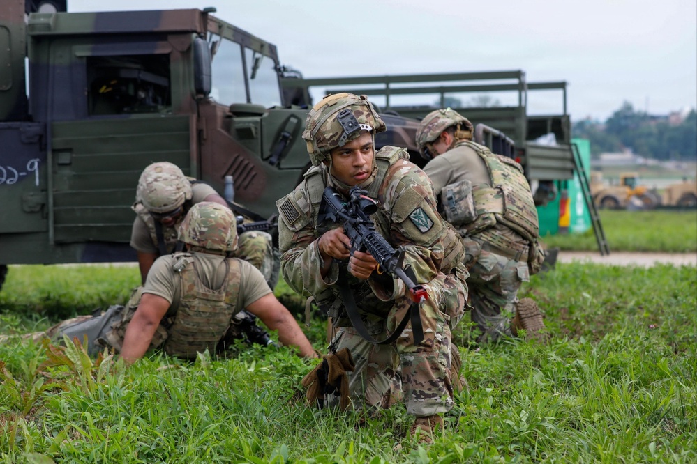 The image shows a group of soldiers in military camouflage (OCP/MultiCam pattern) conducting what appears to be a tactical field exercise or training drill. Multiple soldiers are positioned in low stances near military vehicles, with one soldier in the foreground holding an M4-style rifle and looking directly at the camera. The setting appears to be a grassy field adjacent to a military installation or airfield.