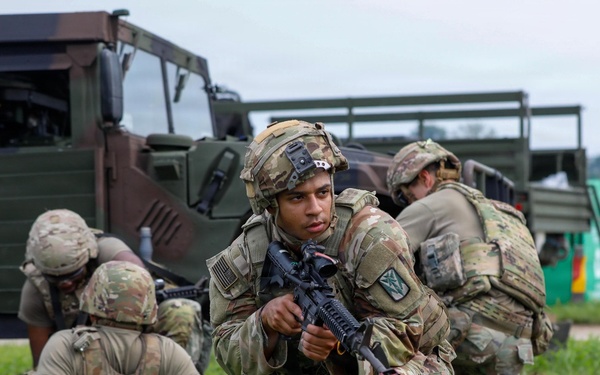 The image shows a group of soldiers in military camouflage (OCP/MultiCam pattern) conducting what appears to be a tactical field exercise or training drill. Multiple soldiers are positioned in low stances near military vehicles, with one soldier in the foreground holding an M4-style rifle and looking directly at the camera. The setting appears to be a grassy field adjacent to a military installation or airfield.