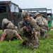 The image shows a group of soldiers in military camouflage (OCP/MultiCam pattern) conducting what appears to be a tactical field exercise or training drill. Multiple soldiers are positioned in low stances near military vehicles, with one soldier in the foreground holding an M4-style rifle and looking directly at the camera. The setting appears to be a grassy field adjacent to a military installation or airfield.