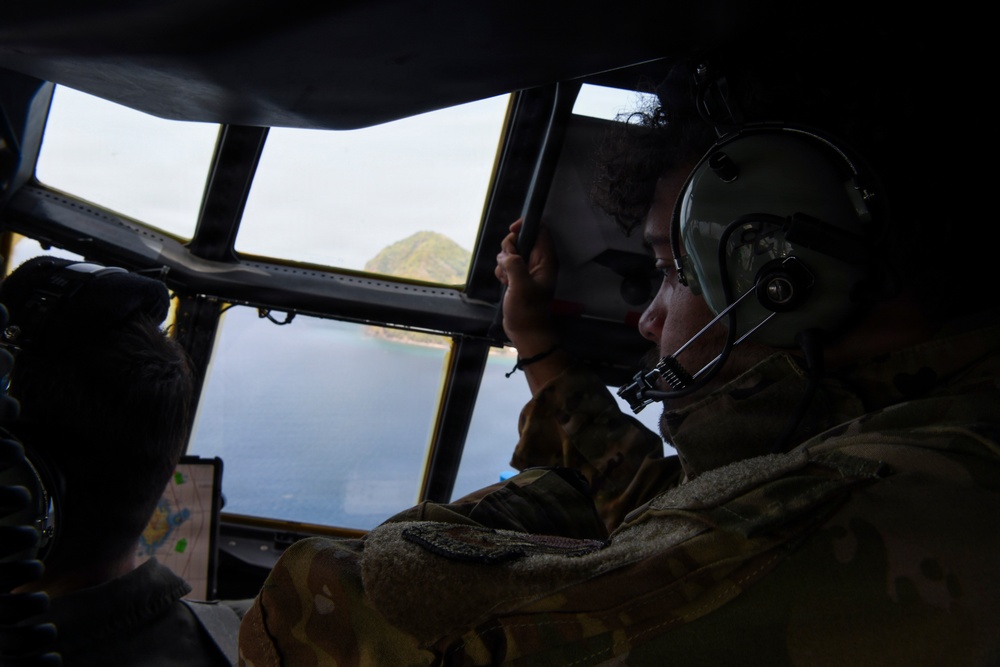 Military aircrew members are visible inside the cockpit of a large military aircraft, likely a transport or patrol aircraft, flying over a coastal area with a rocky island or promontory visible through the windscreen. At least two personnel in camouflage uniforms and flight helmets with communication headsets are observing the terrain below. A navigation display or tablet is visible on the instrument panel.