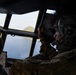 Military aircrew members are visible inside the cockpit of a large military aircraft, likely a transport or patrol aircraft, flying over a coastal area with a rocky island or promontory visible through the windscreen. At least two personnel in camouflage uniforms and flight helmets with communication headsets are observing the terrain below. A navigation display or tablet is visible on the instrument panel.