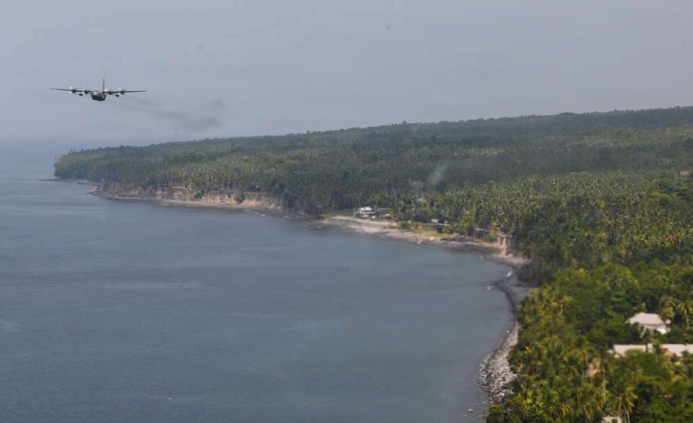A military transport aircraft, appearing to be a C-130 Hercules, flies at low altitude over a tropical coastline. The coastline features dense palm tree vegetation, rocky cliffs, and a small coastal settlement visible along the shoreline. The image is taken from an elevated aerial perspective, possibly from another aircraft.